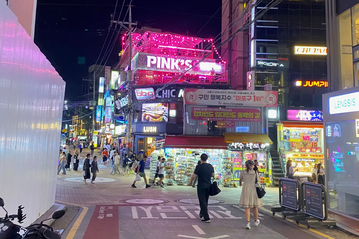 ถนนในฮงแดยามค่ำคืน มีป้ายไฟนีออน ไฟประดับบนดาดฟ้า และผู้คนเดินผ่านร้านเล็กๆ